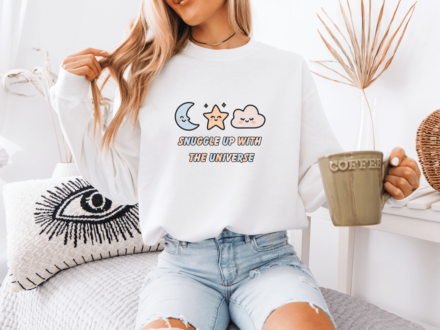 Woman wearing a white “Snuggle Up With the Universe” sweatshirt, holding a coffee mug and relaxing on a cozy bed