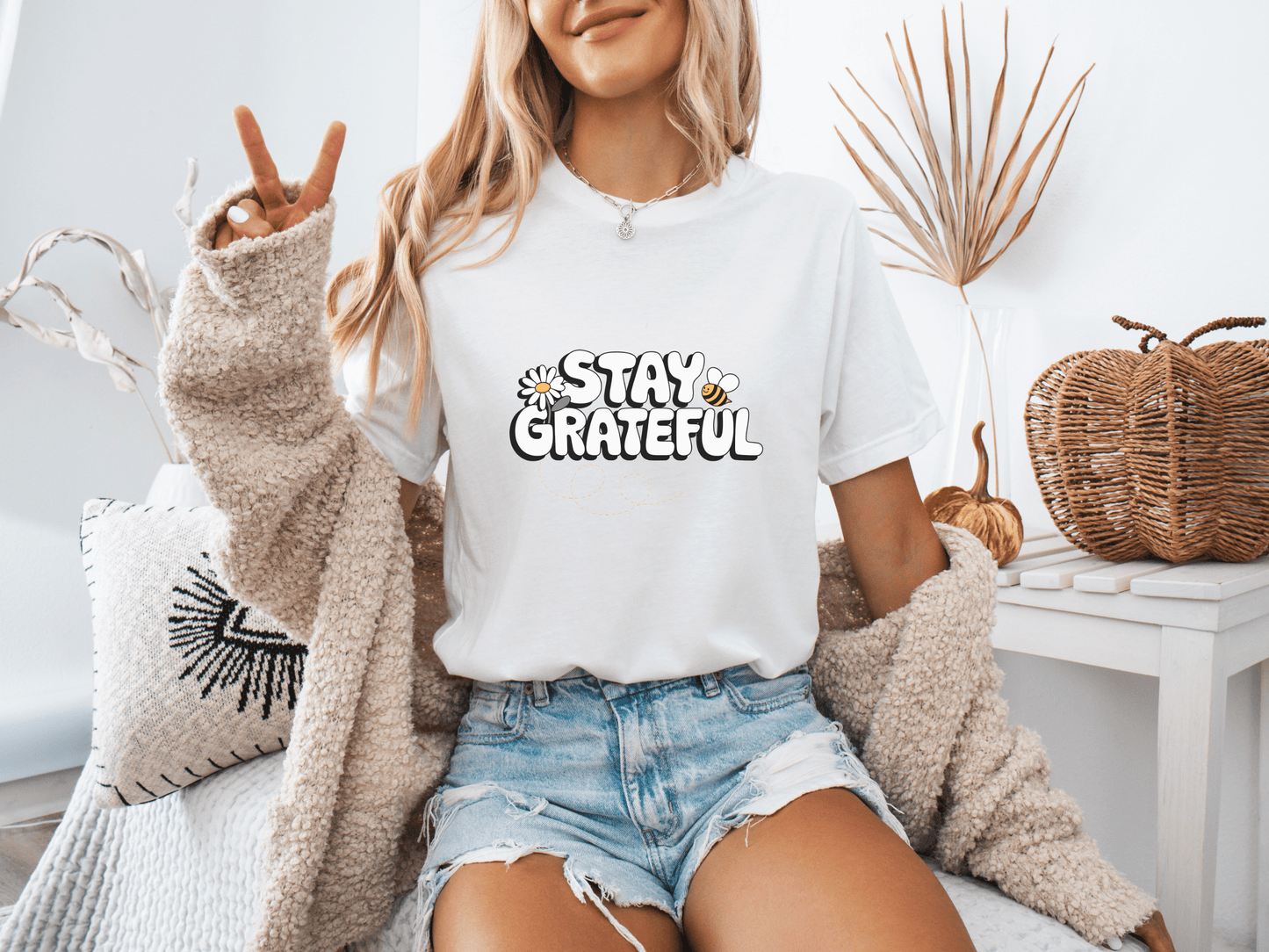 Woman wearing a white “Stay Grateful” t-shirt with playful daisy accents, posing in a cozy boho room