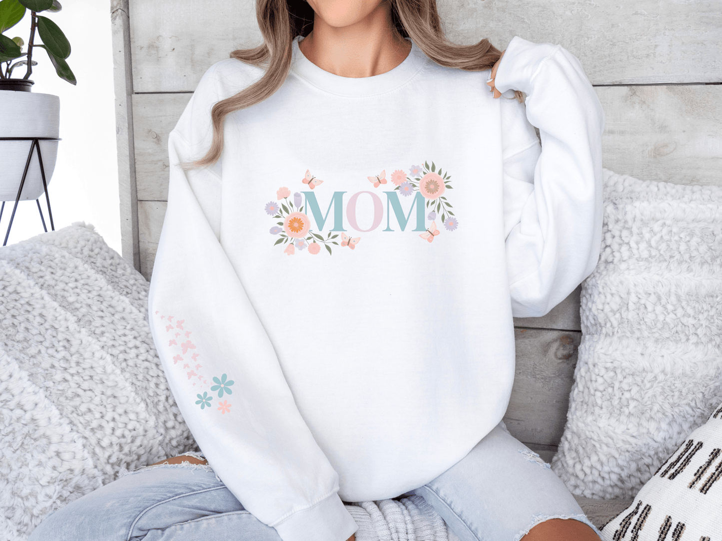 Woman wearing a white sweatshirt with pastel floral “MOM” lettering, seated on a cozy couch with textured pillows