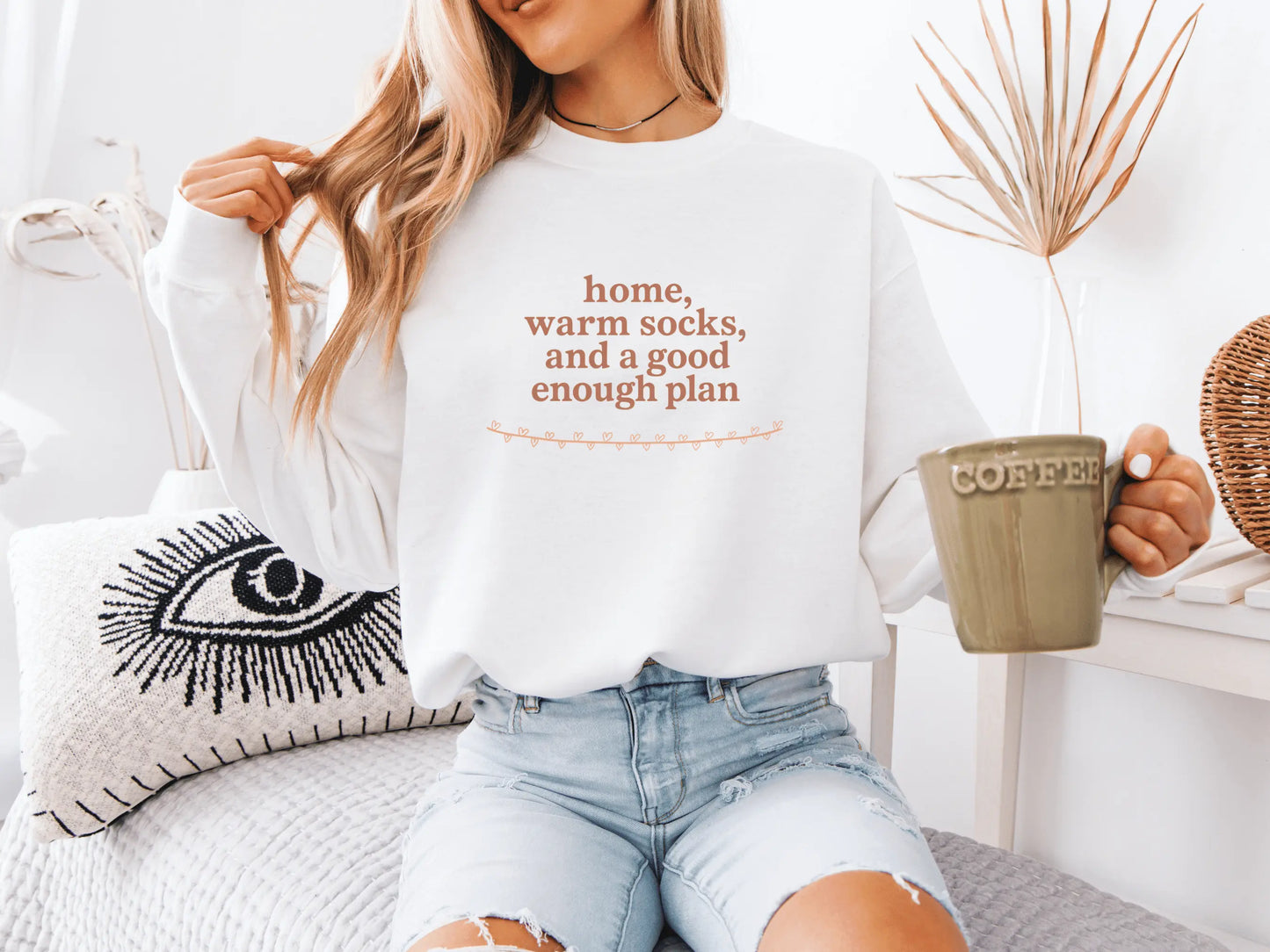 Person wearing a white sweatshirt with text "home, warm socks, and a good enough plan", holding a mug, sitting on a couch.
