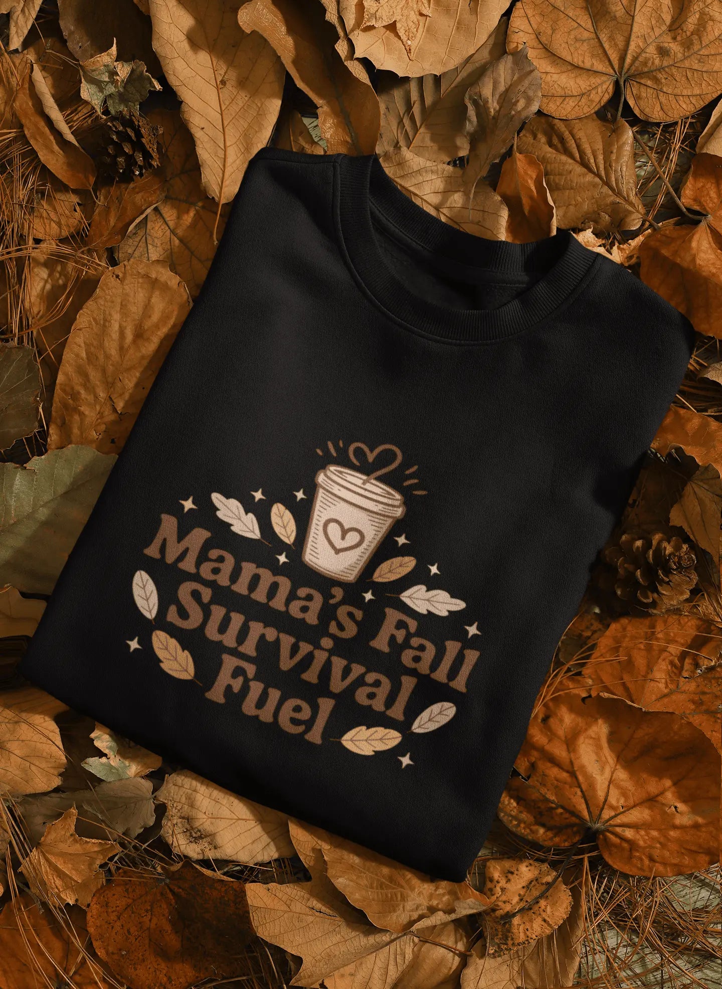 Black sweatshirt with 'Mama's Fall Survival Fuel' design on a bed of autumn leaves.