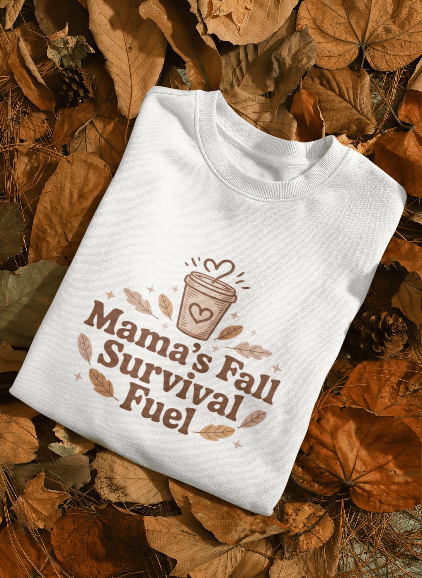 White sweatshirt with 'Mama's Fall Survival Fuel' text and graphic on a bed of autumn leaves.