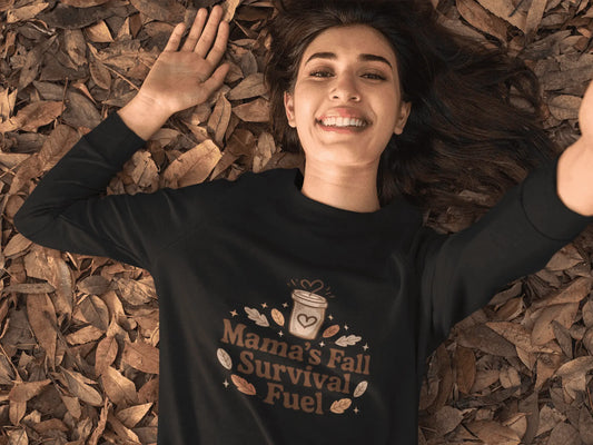 Person wearing a black sweatshirt with 'Mama's Fall Survival Fuel' text, lying on fall leaves.
