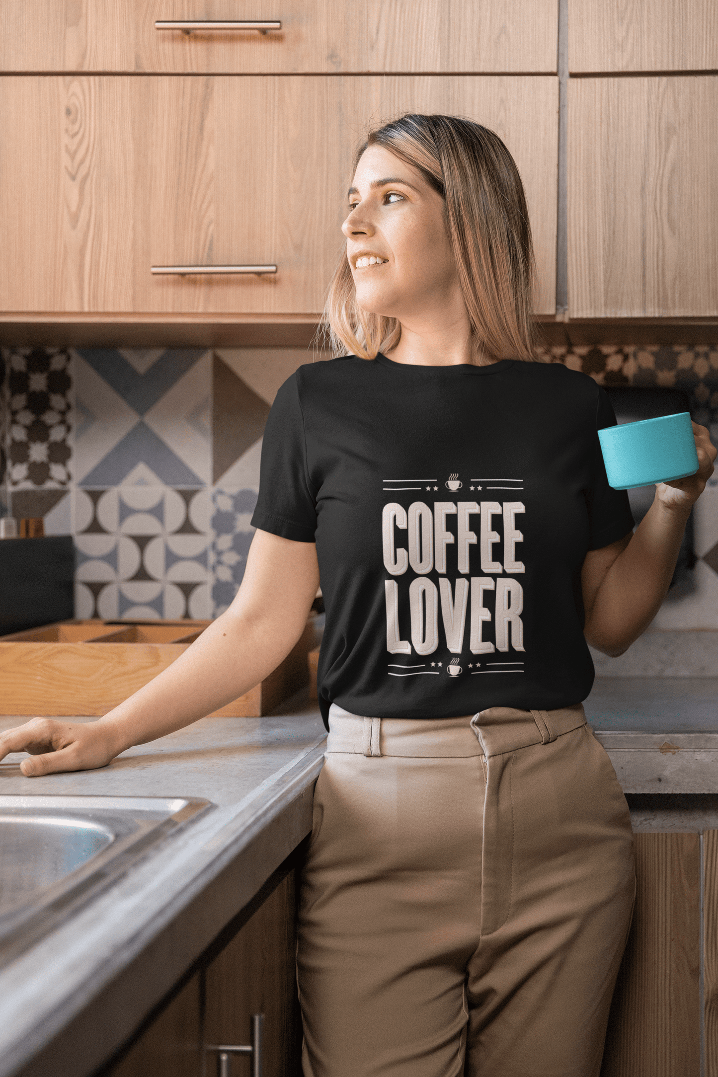 Smiling woman in kitchen wearing a black "Coffee Lover" t-shirt while holding a turquoise mug — cozy morning vibes for caffeine fans.