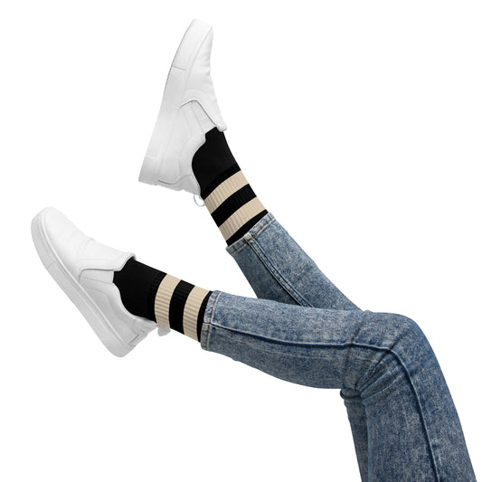 Person wearing black and beige striped coffee-themed socks styled with blue jeans and white slip-on sneakers, legs playfully raised against a white background.