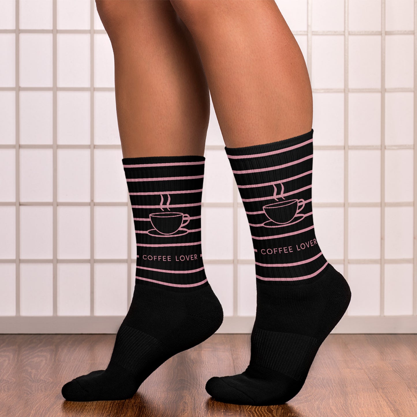 Reverse view of person wearing “Coffee Lover” socks with pink coffee cup and line art, captured against a tiled wall and wooden floor — cozy and bold for coffee fans.