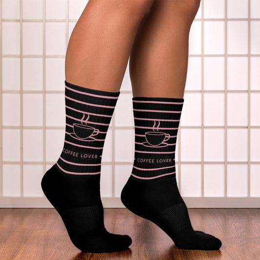 Person wearing black sublimated “Coffee Lover” socks with pink lines and coffee cup design, standing on wooden floor in front of tiled wall — stylish brew-themed comfort for your feet.
