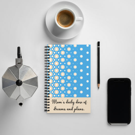 Blue notebook with white and yellow stars and inspirational quote –Mom's daily dose of dreams and plans- front cover view on cozy desk setup