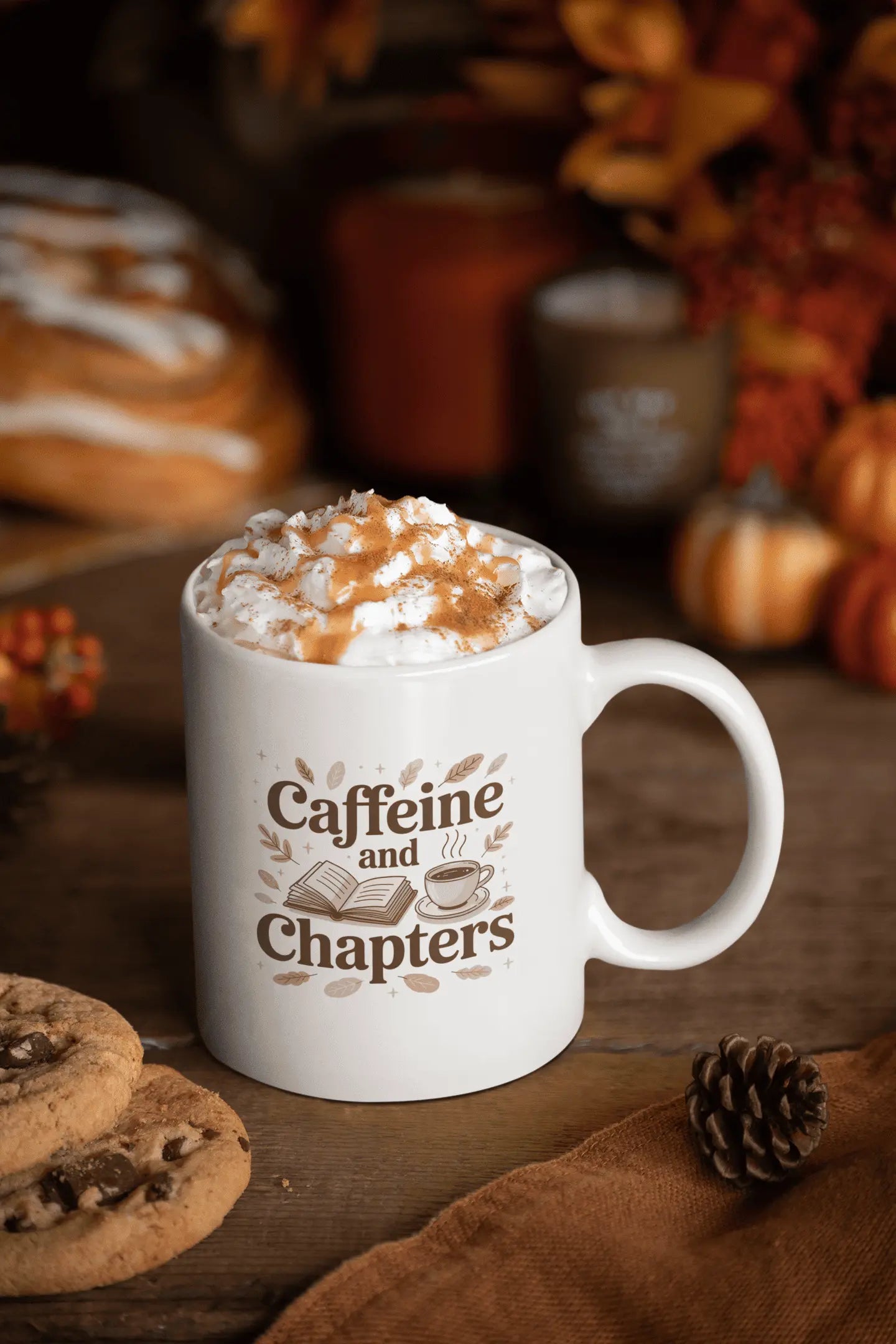 White mug with 'Caffeine and Chapters' text, filled with hot chocolate, surrounded by fall-themed items.