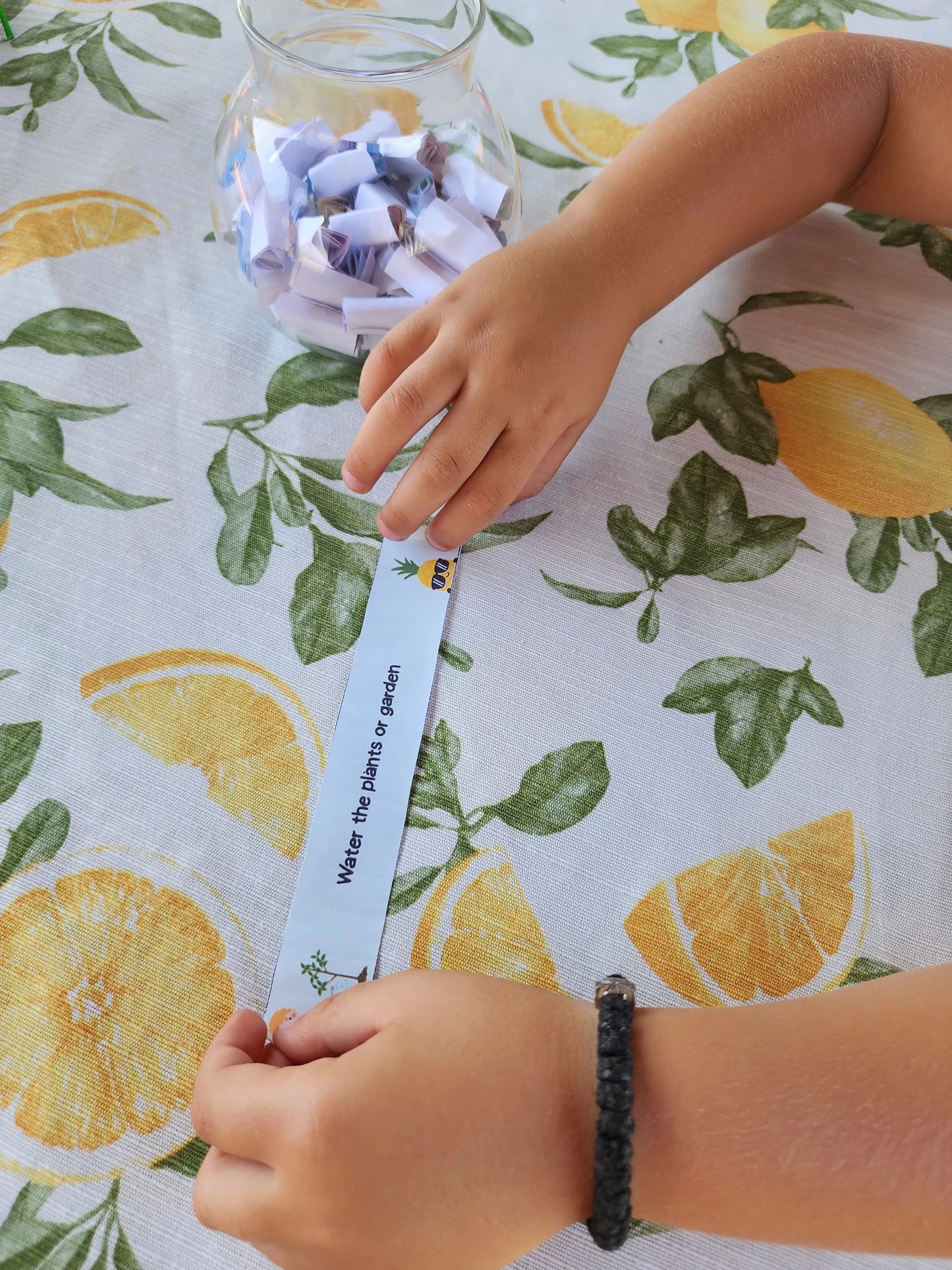 A child holding a printable activity strip that says "Water the plants or garden" next to a full activity jar on a citrus-themed tablecloth.