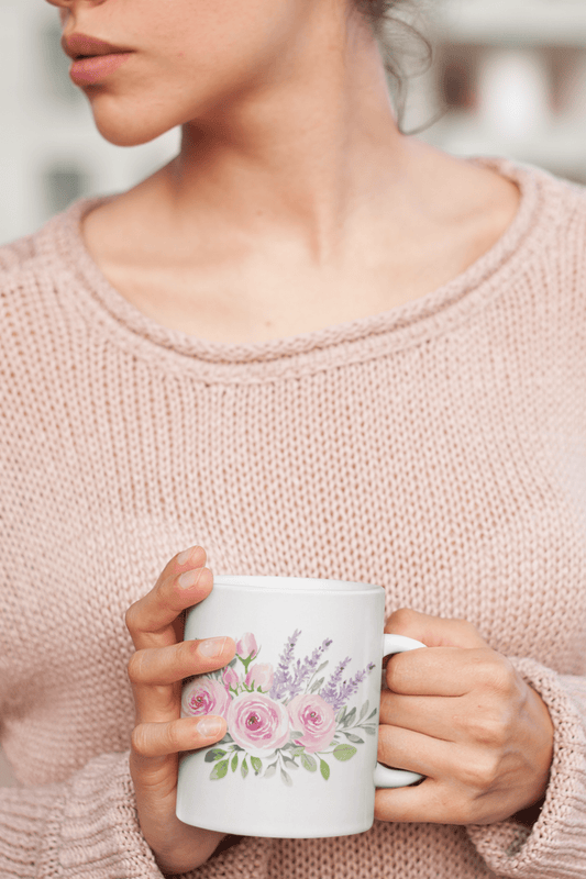 Woman in a pink sweater holding a white mug with a soft pink and lavender floral bouquet, close-up on hands and chest.