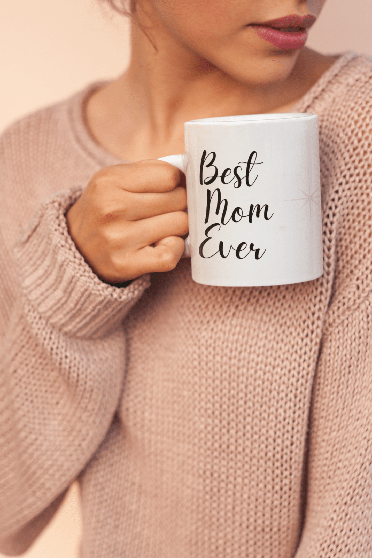 Woman in a cozy pink sweater holding a white mug that says “Best Mom Ever,” close-up of hands and lips.