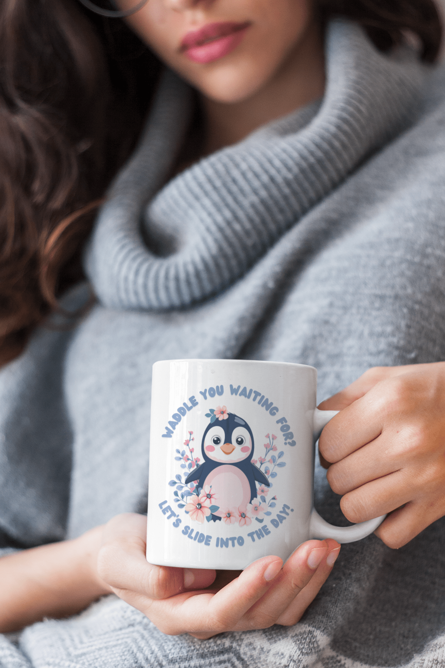 A woman in a gray sweater holding a white mug with a cute penguin design and the text "Waddle You Waiting For? Let’s Slide Into the Day!" surrounded by pink and blue flowers.