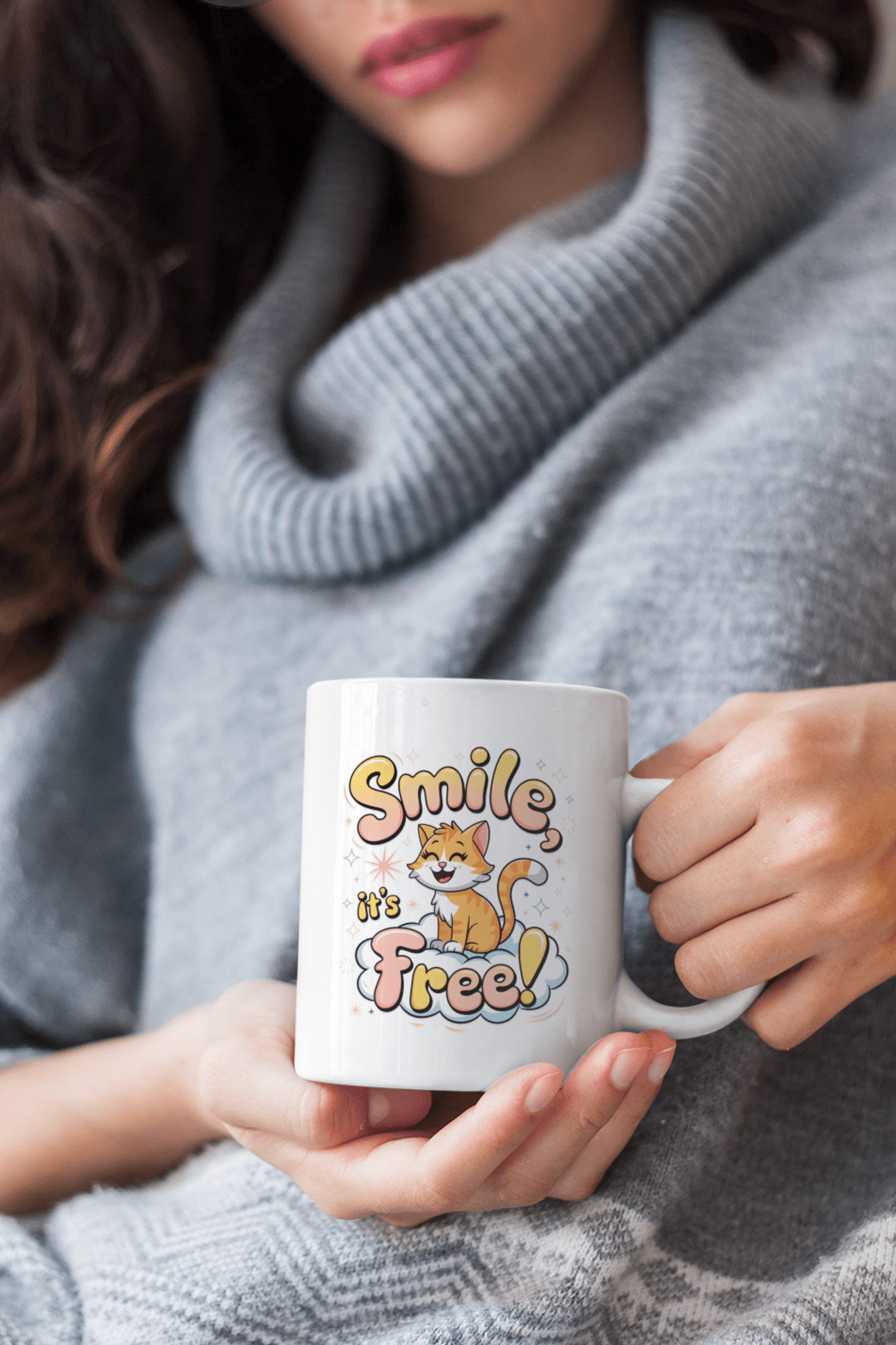 Woman in a cozy gray sweater holding a white mug with a cheerful cat and the phrase “Smile, it’s Free!” in playful, colorful text.