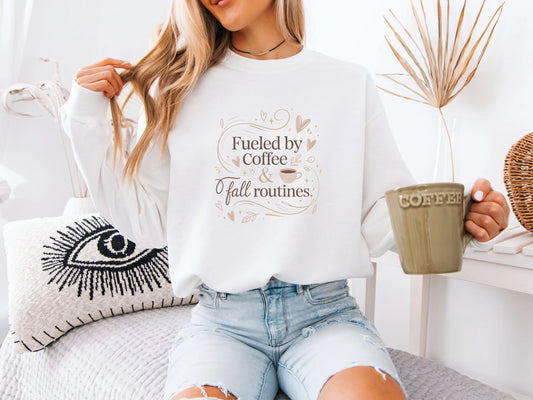 Woman wearing a white sweatshirt with a coffee-themed design and text "Fueled by coffee & fall routines" holding a mug, sitting on a couch