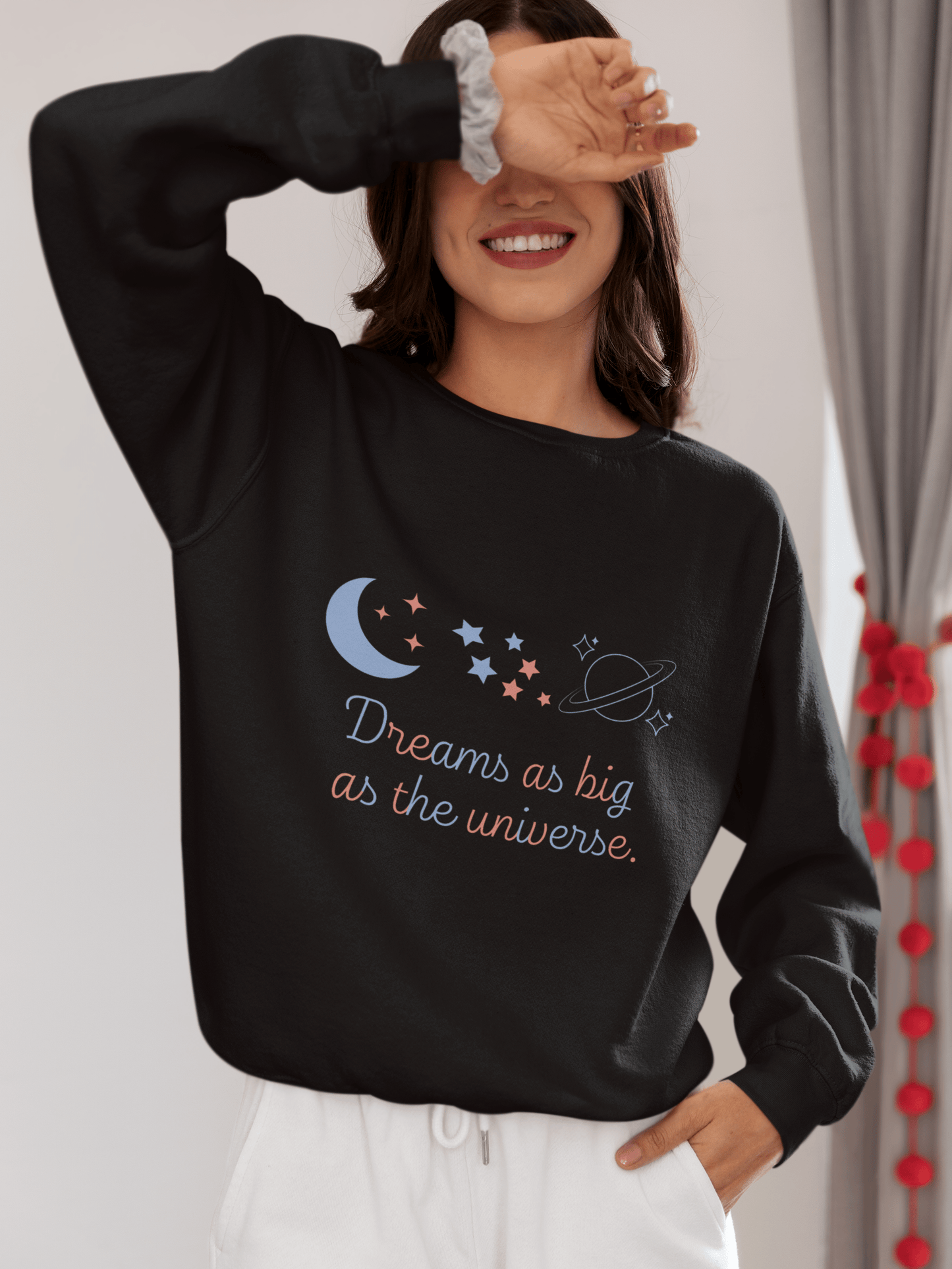 Smiling woman wearing a black sweatshirt with pastel-colored celestial elements and the quote “Dreams as big as the universe”; she’s playfully covering her eyes with one hand, standing in a cozy indoor setting.