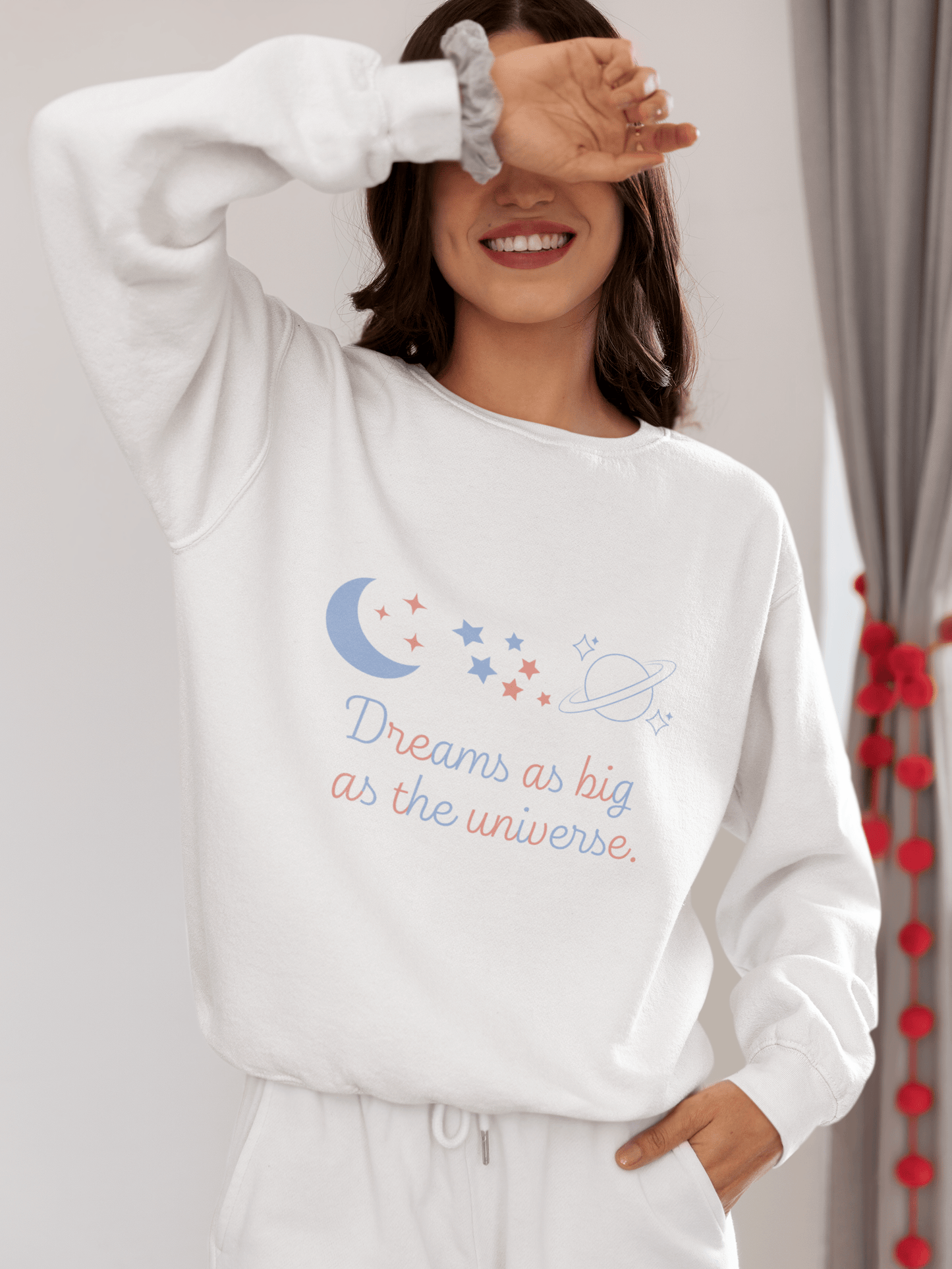 Smiling woman wearing a white sweatshirt with pastel-colored celestial elements and the quote “Dreams as big as the universe”; she’s playfully covering her eyes with one hand, standing in a cozy indoor setting.
