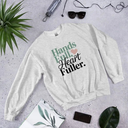 White sweatshirt with 'Hands Full, Heart Fuller' text on a gray surface with accessories.