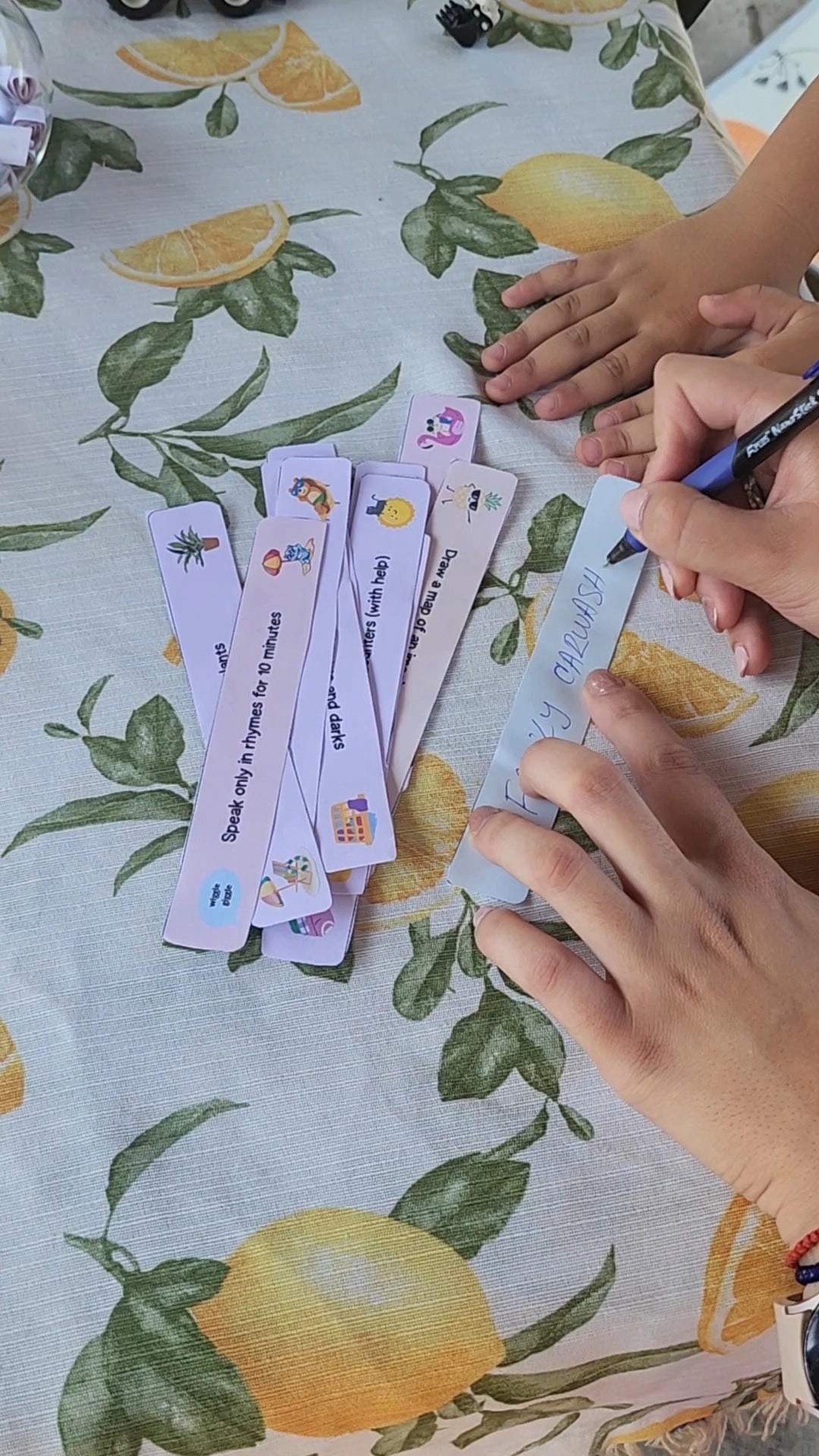 Child’s hands writing a custom summer activity on a blank printable strip labeled “Familly Carwash,” surrounded by colorful prompt cards on a lemon-patterned tablecloth