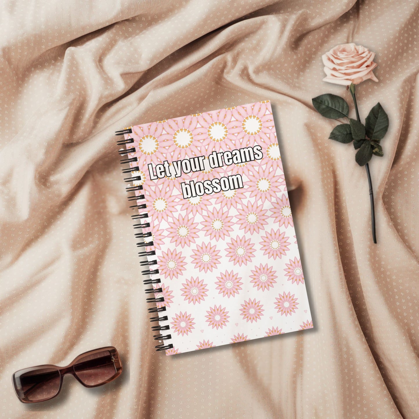 Spiral notebook with “Let your dreams blossom” quote on pink floral cover – styled on soft fabric with rose and sunglasses