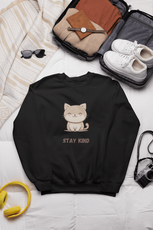 Black sweatshirt with a cute cat and the words “Stay Kind” placed on a bed, next to a travel suitcase, sneakers, folded clothes, sunglasses, and travel accessories.