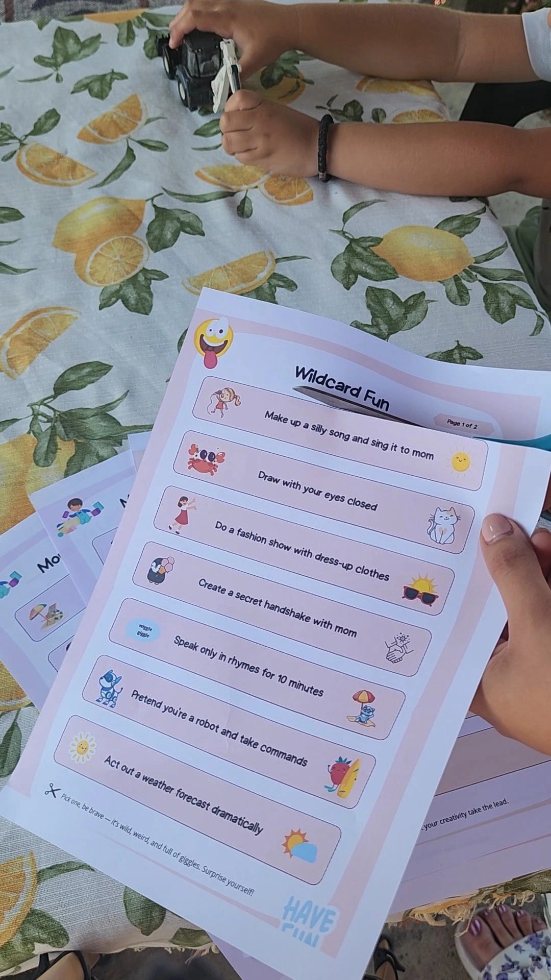 Cut-out paper strips with summer activities for kids, laid out on a table beside a child's hands