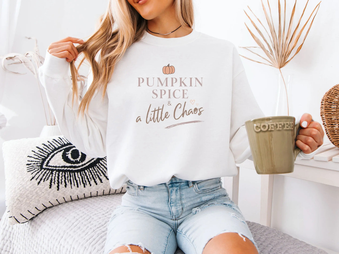 Person wearing a white sweatshirt with 'Pumpkin Spice a Little Chaos' text, holding a mug, sitting on a couch.