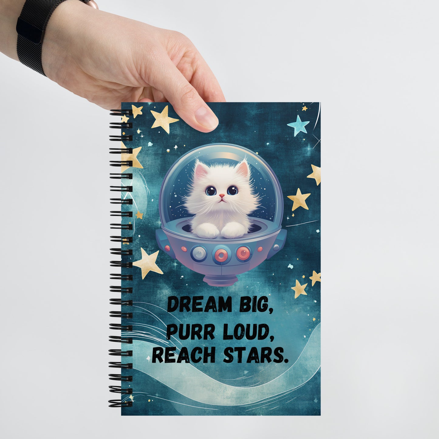 A hand holding a spiral notebook with a blue galaxy-themed cover, featuring a white cat in a space helmet surrounded by stars. The cover text reads: “Dream Big, Purr Loud, Reach Stars.”