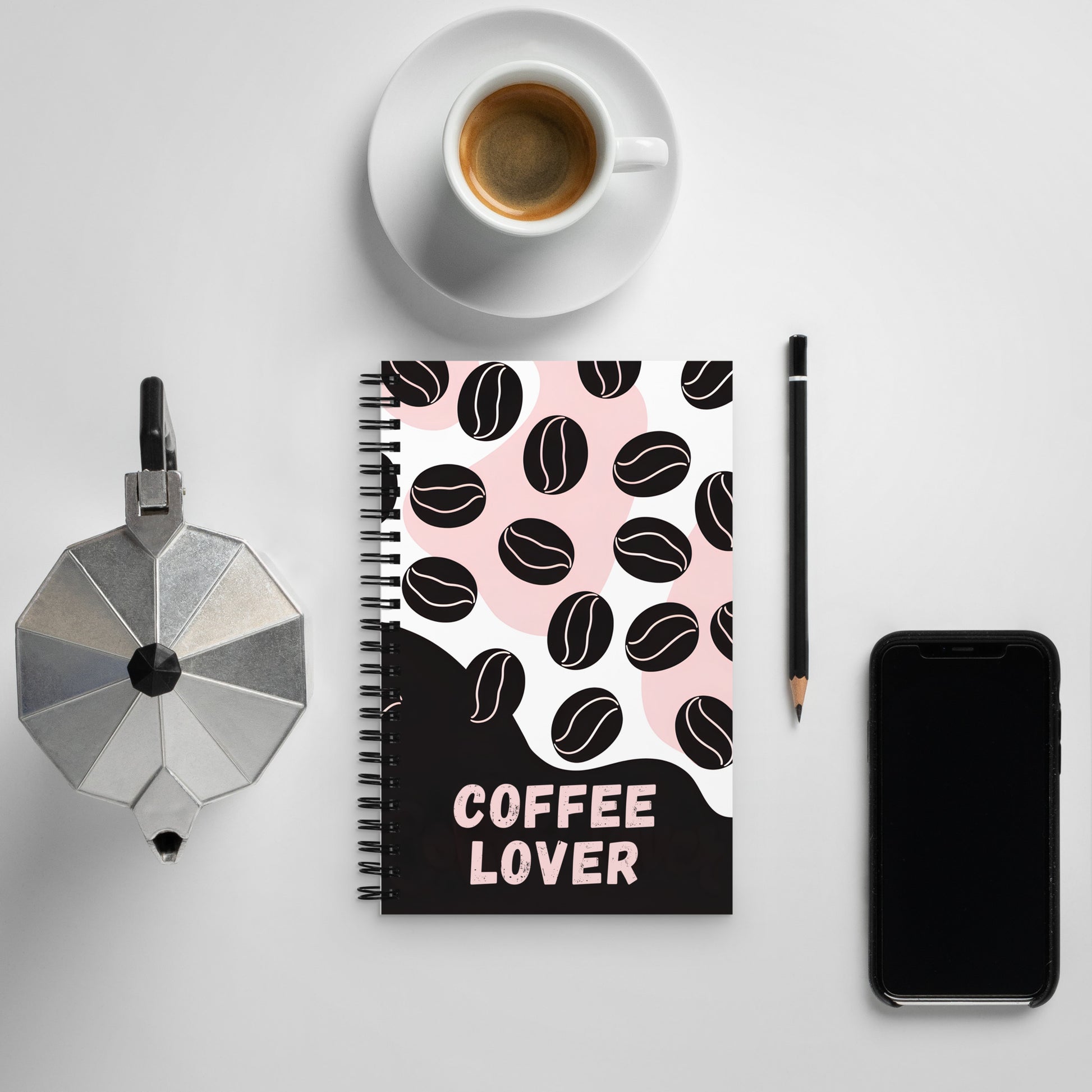 Flat lay of a “Coffee Lover” spiral notebook with black and pink coffee bean design, placed on a white desk alongside an espresso cup, moka pot, pencil, and smartphone — the perfect setup for your next brew-fueled brainstorm.