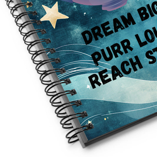 Close-up of a spiral-bound notebook with a blue cosmic-themed cover, showing the phrase “Dream Big, Purr Loud, Reach Stars” and black wire binding.
