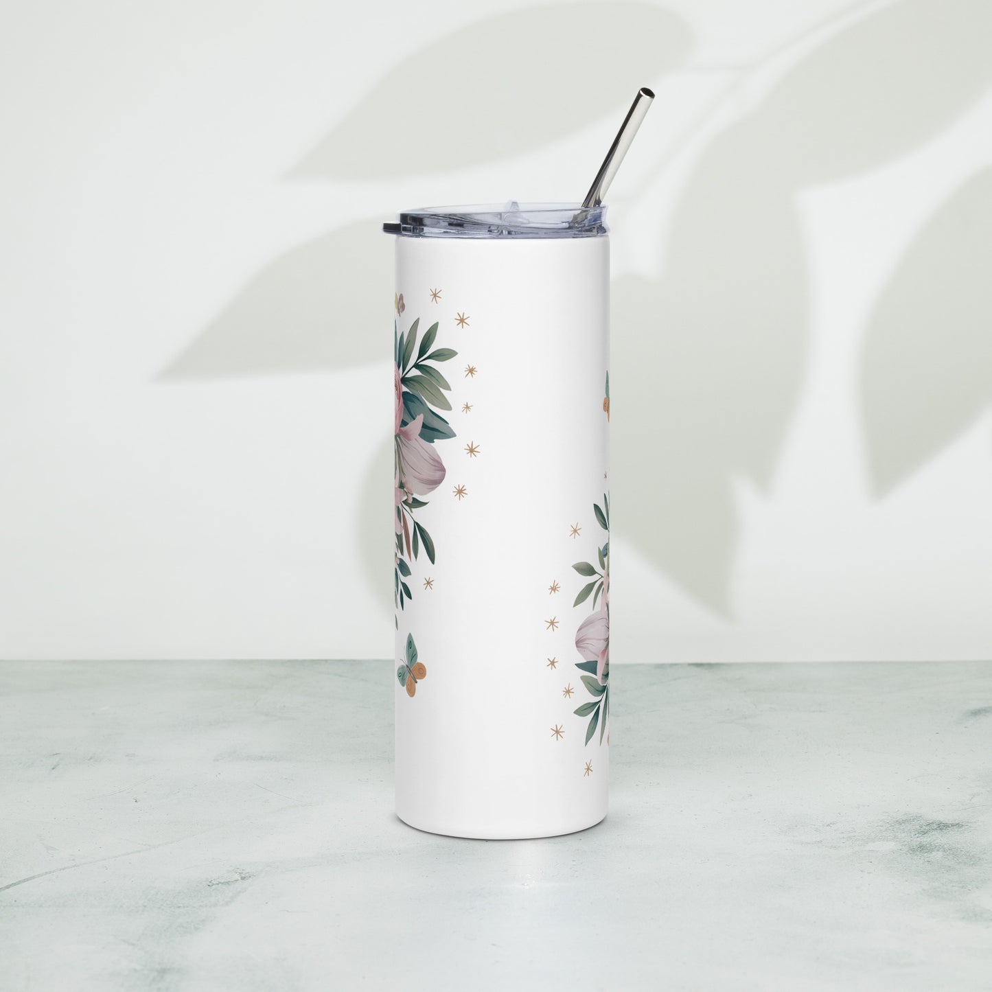 White stainless steel tumbler with partial pink floral design and butterflies on the back side, standing upright on a light green surface with soft leaf shadows on the wall.