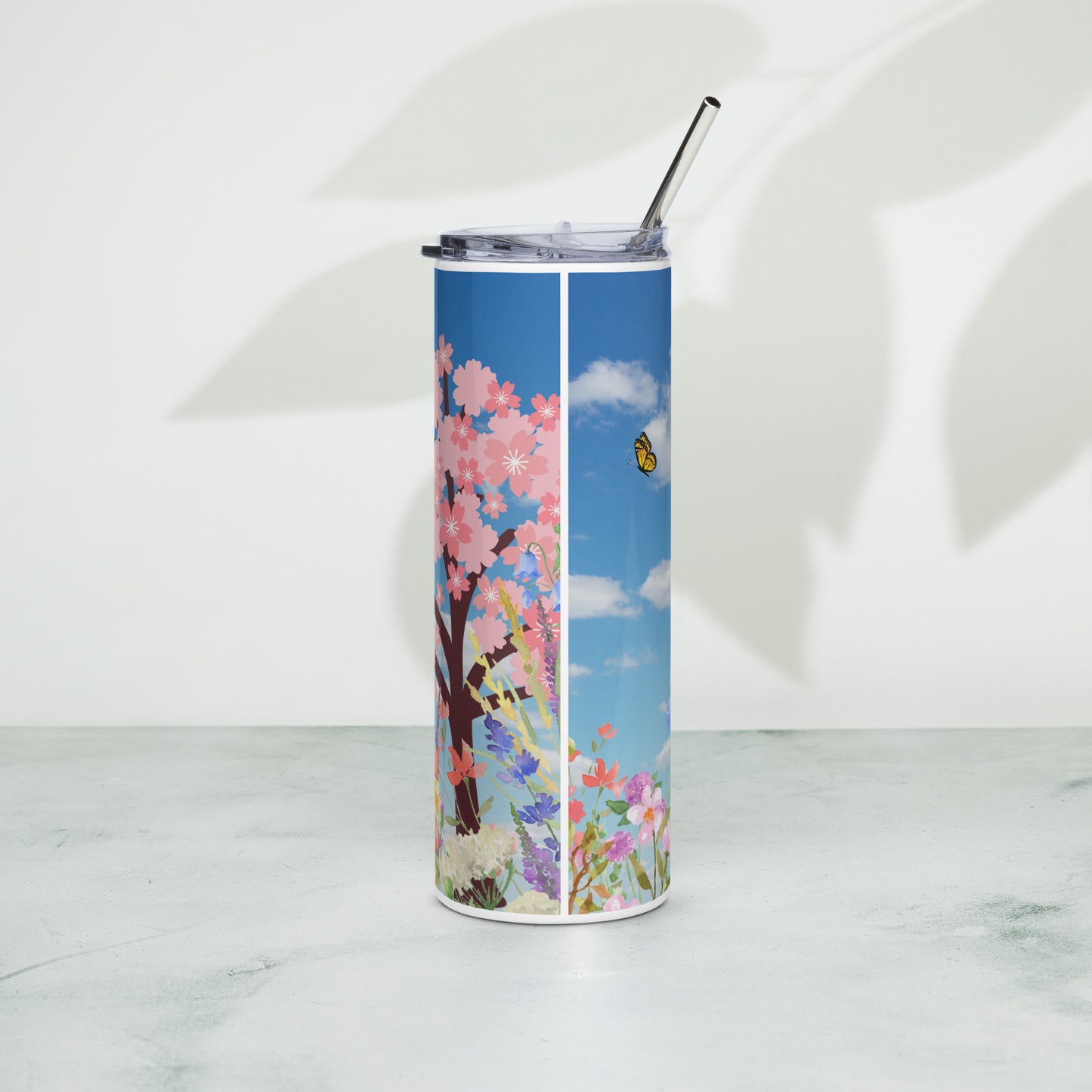 Tall stainless steel tumbler with a metal straw, decorated with a spring floral pattern and butterfly on a blue sky background, standing upright on a white surface with soft shadows in the background.