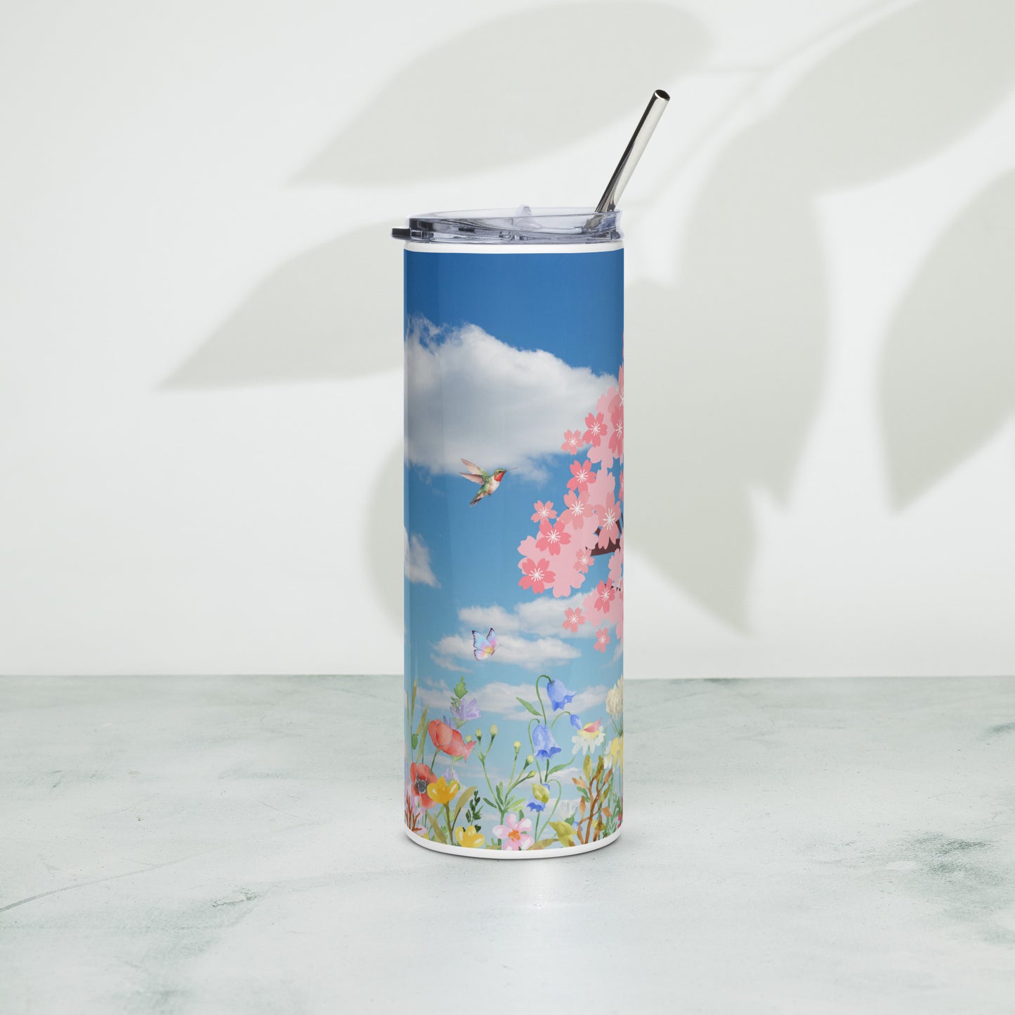 Stainless steel tumbler with a metal straw, featuring a bright blue sky with clouds, cherry blossoms, wildflowers, and butterflies, standing upright on a light surface with soft leaf shadows in the background.