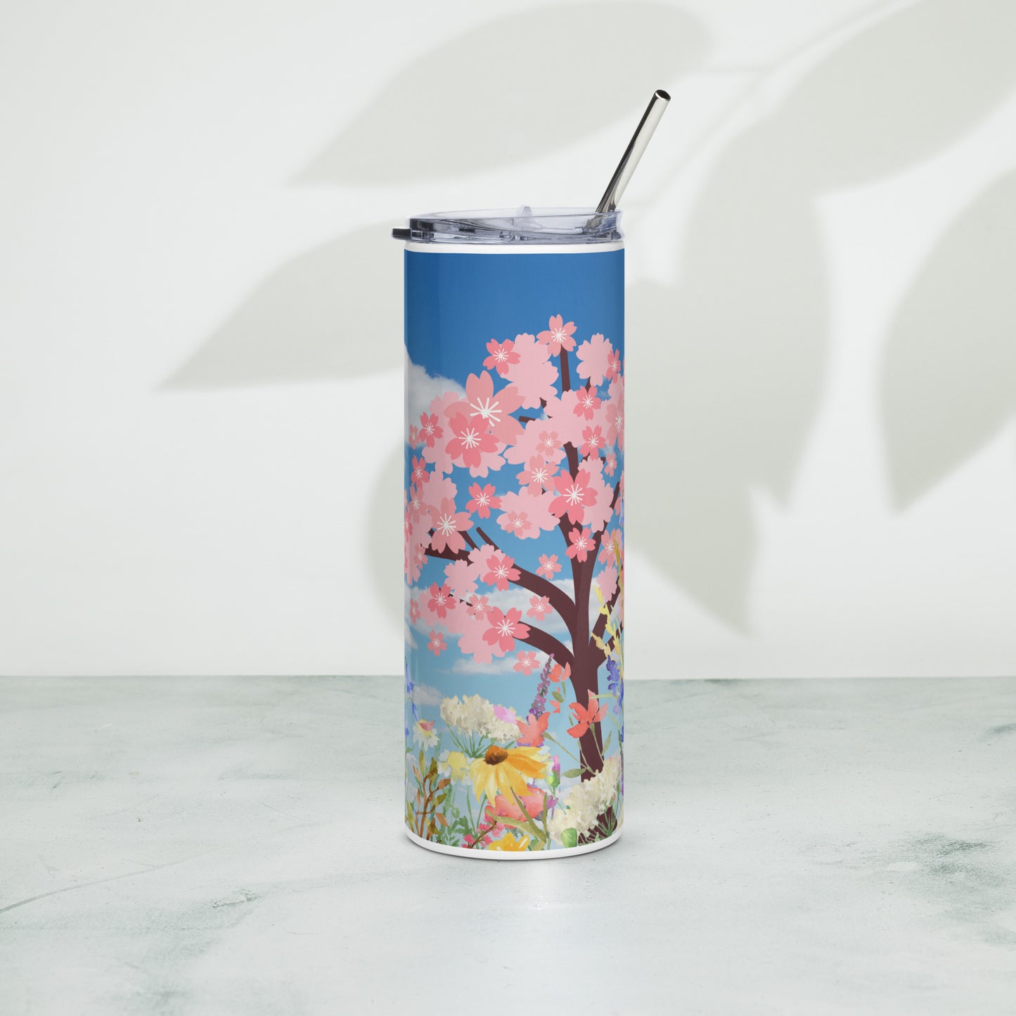 Stainless steel tumbler with metal straw, featuring a vivid cherry blossom tree and pastel floral design on a blue sky background, placed upright on a light surface.