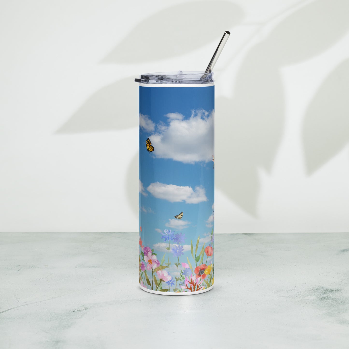 Stainless steel tumbler with a metal straw, decorated with blue sky, fluffy white clouds, butterflies, and colorful spring flowers, standing on a light surface with soft leaf shadows in the background.