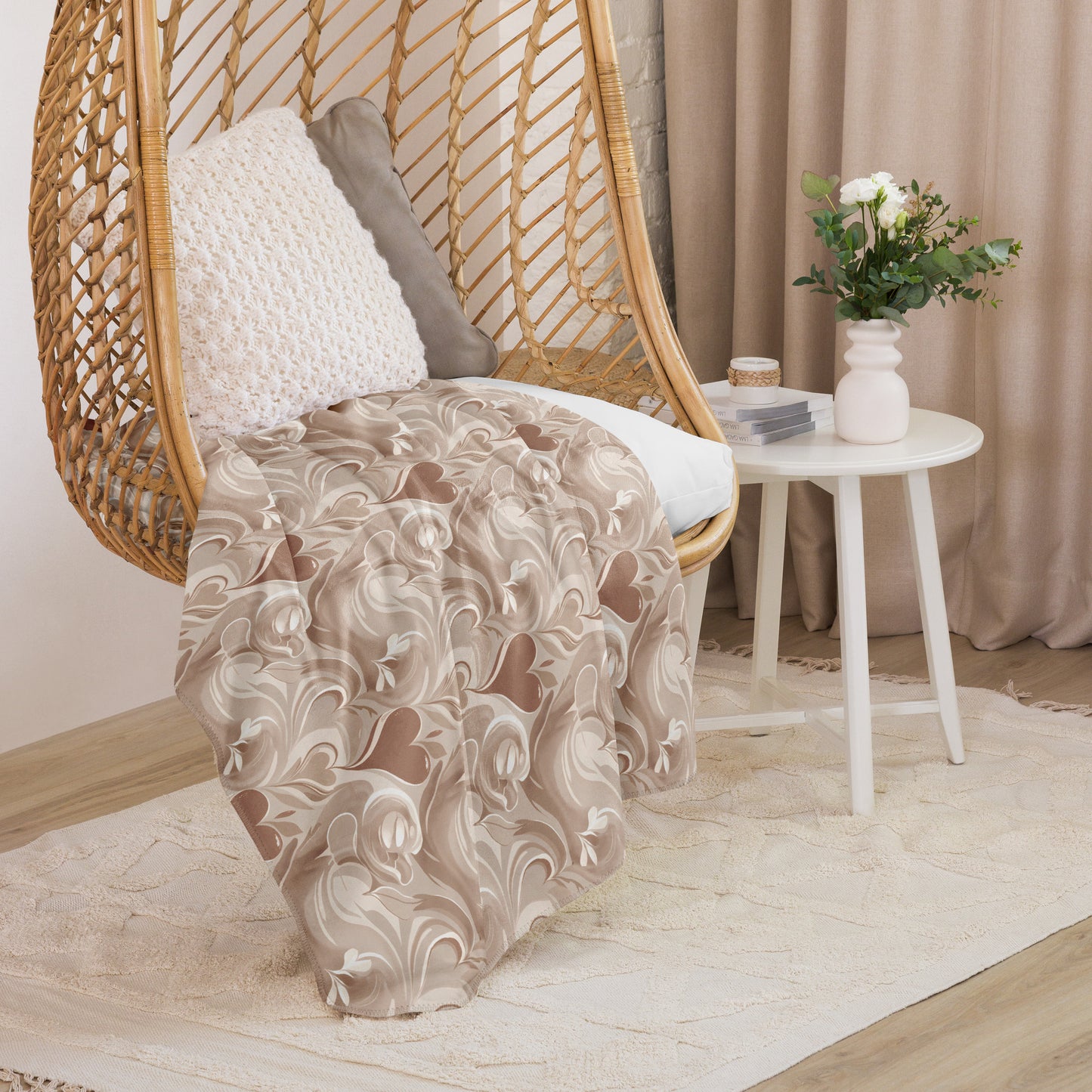 Tan sherpa blanket with cream coffee-inspired swirl design draped over a wicker hanging chair with decorative pillows, next to a white round side table with flowers and books in a soft, neutral-toned reading nook.