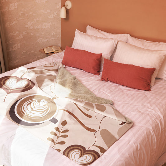 Tan and brown sherpa blanket featuring large latte art and coffee bean illustrations, styled on a blush pink bed with terracotta and white pillows in a sunlit bedroom with soft, neutral decor.