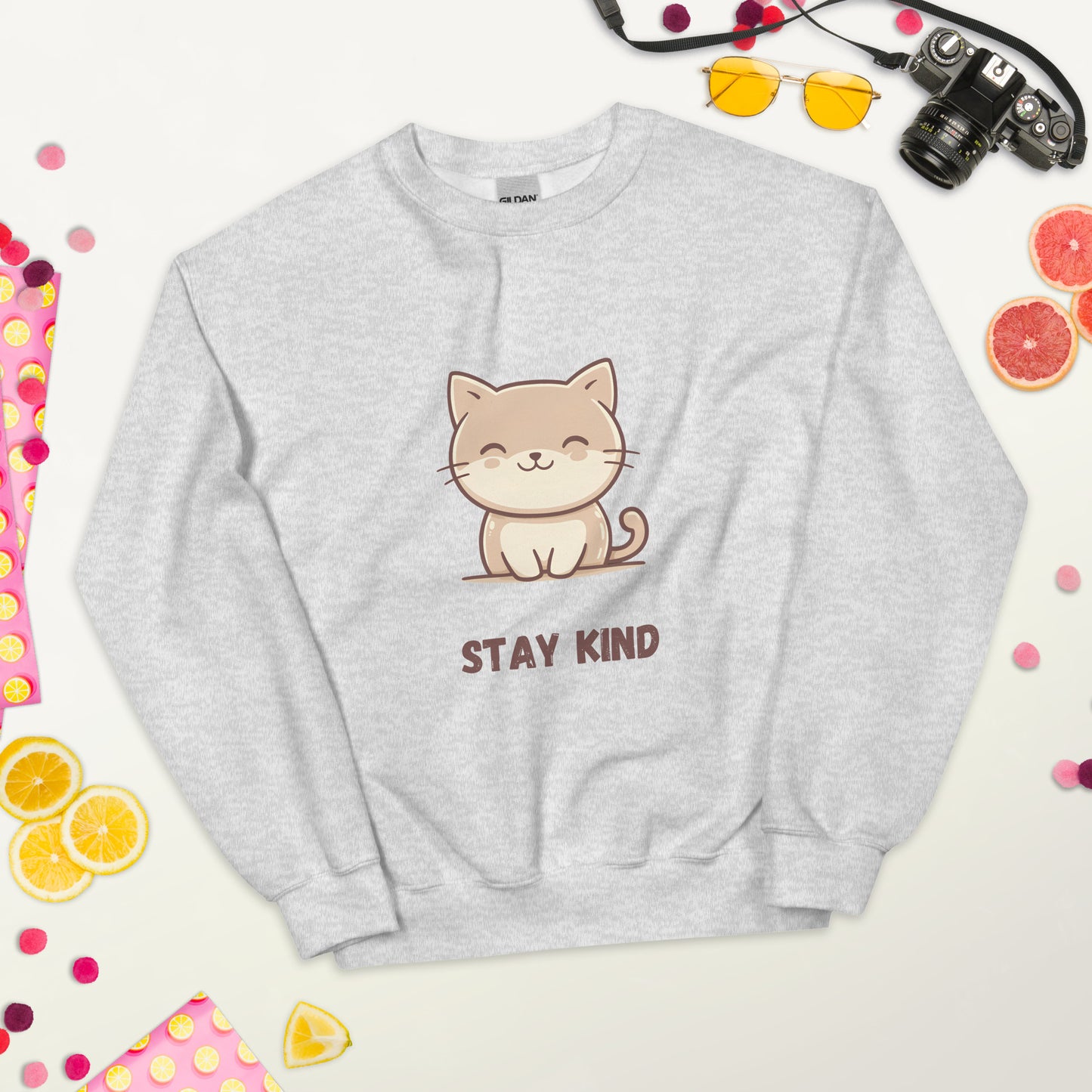 Ash gray sweatshirt with a cute cat and the phrase “Stay Kind,” laid flat on a white surface, surrounded by colorful fruits, sunglasses, a camera, and playful decorative elements.