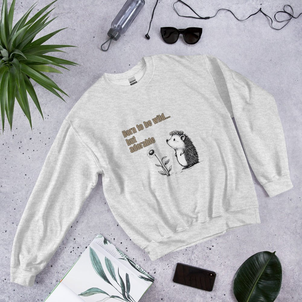 Ash gray sweatshirt with a cute hedgehog and “Born to be wild… but adorable” text, styled flat lay with sunglasses, phone, and green leaves.