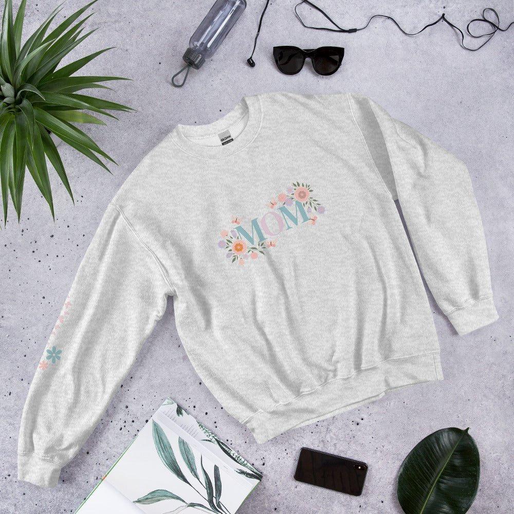 Light gray sweatshirt with pastel floral “MOM” lettering, laid flat on a cozy lifestyle scene with sunglasses, plants, phone, and journal – stylish gift idea for moms.