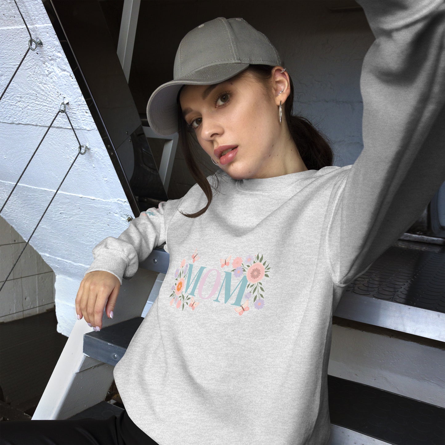 Young woman in a gray baseball cap posing in a cozy light gray sweatshirt with pastel floral “MOM” design – casual street-style vibe with a soft, feminine twist.