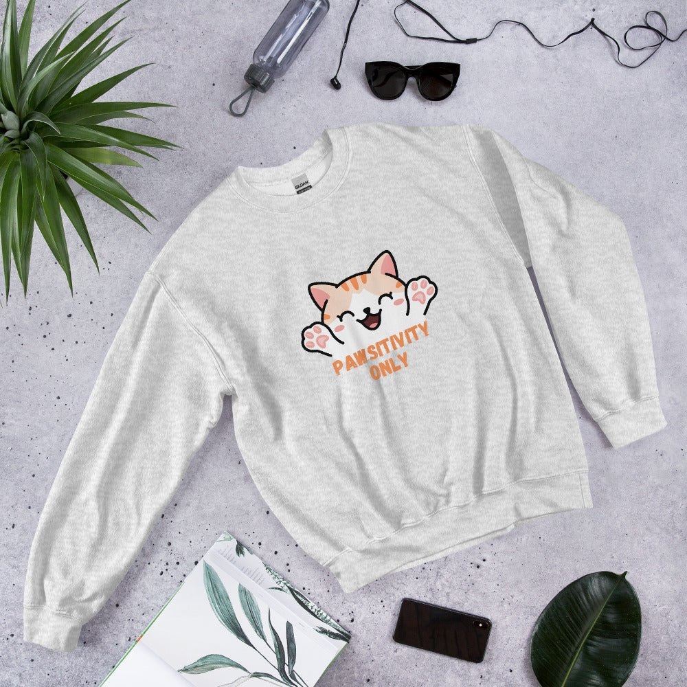 Light grey crewneck sweatshirt with a cute orange cat and “Pawsitivity Only” text, laid flat on a stone surface surrounded by sunglasses, a plant, phone, and notebook.