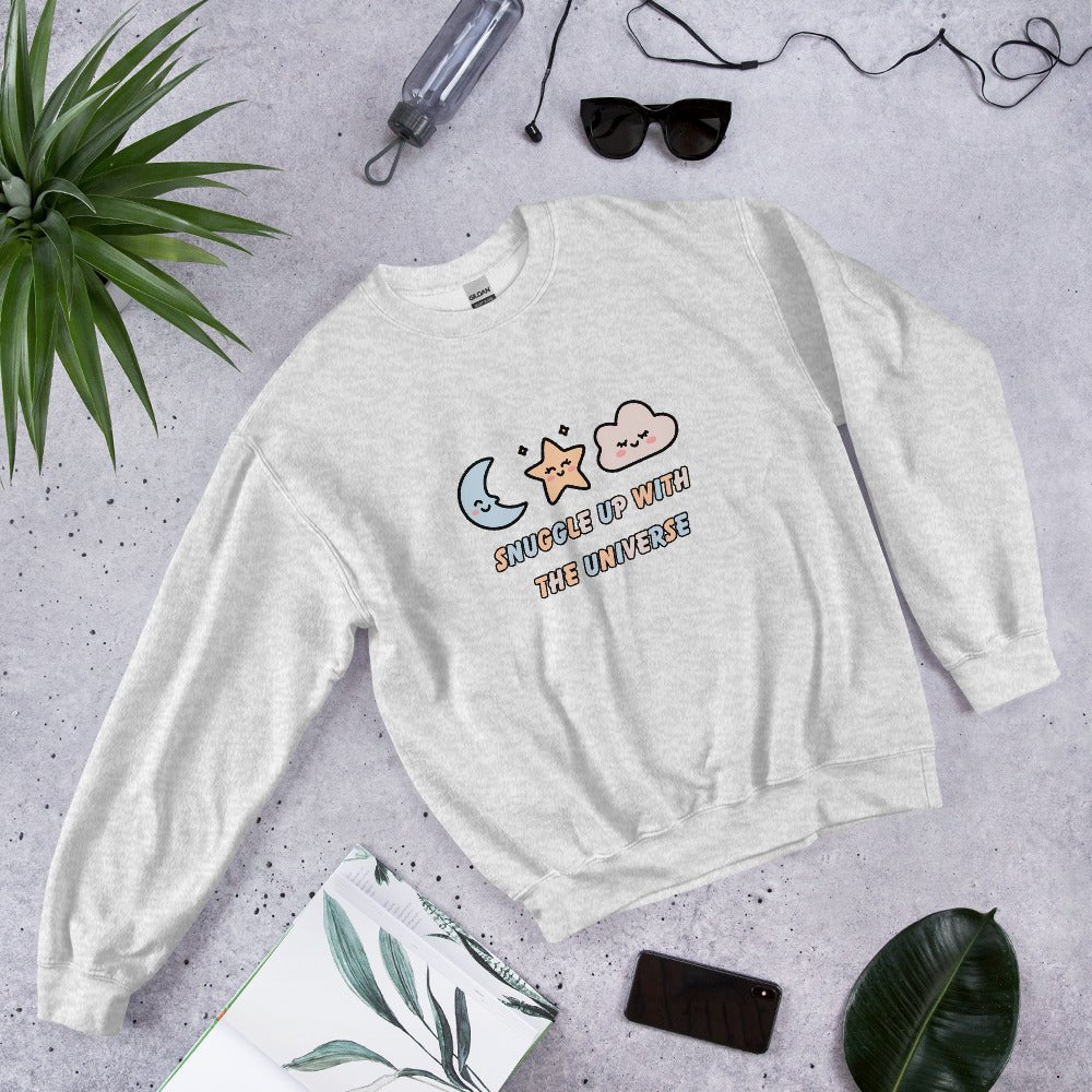Ash gray sweatshirt with the phrase “Snuggle Up with the Universe” and illustrations of a moon, star, and clouds, laid flat on a concrete background next to sunglasses, a water bottle, and tropical leaves.
