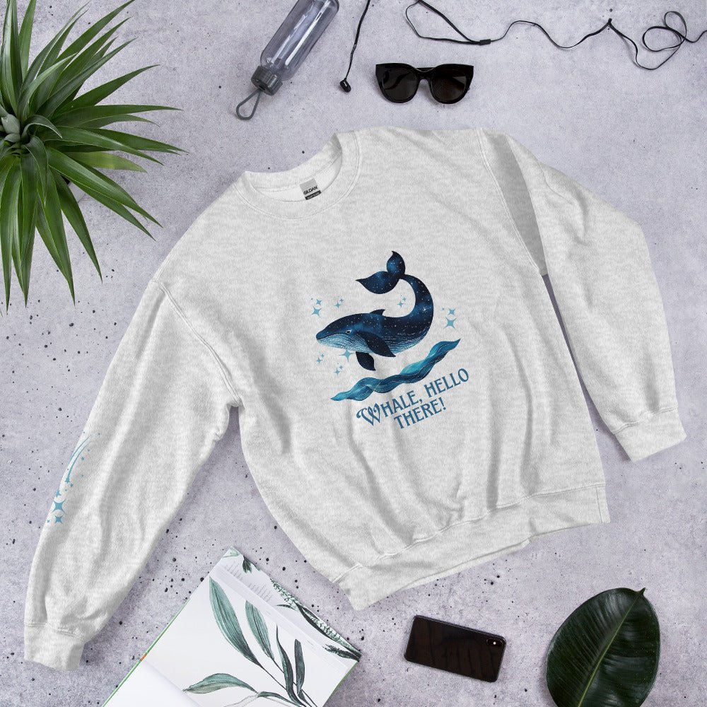 Flat lay of a gray sweatshirt with a cute whale and ocean wave print, reading “Whale, Hello There!” – styled with sunglasses, a water bottle, and tropical leaves on a concrete background.