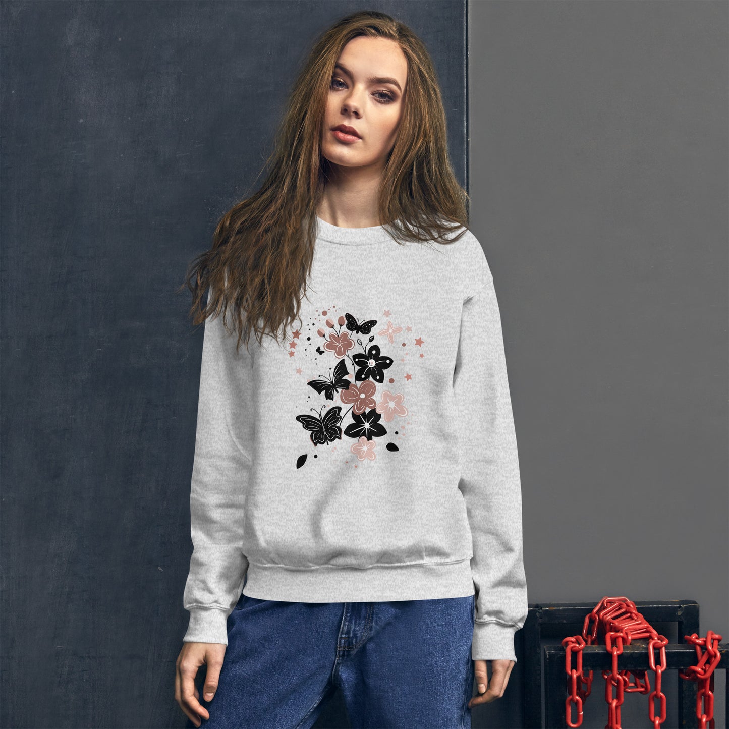 Woman wearing a light gray sweatshirt with a black and pink floral butterfly design on the chest, standing in front of a dark textured wall.