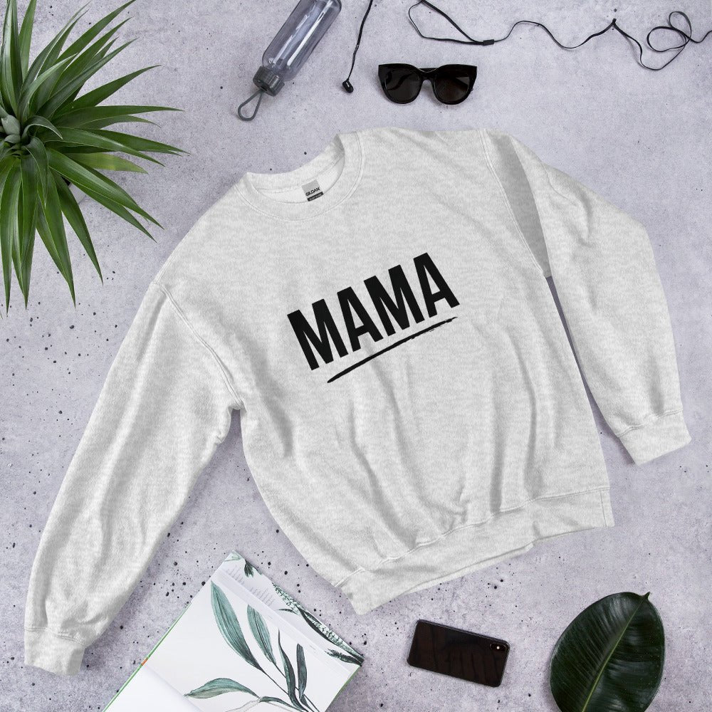 Flat lay of ash grey MAMA sweatshirt with bold black text and underline, surrounded by sunglasses, a plant leaf, notebook, and smartphone