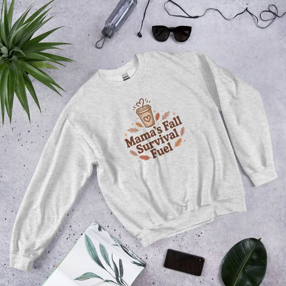 White sweatshirt with 'Mama's Fall Survival Fuel' text and graphic on a gray surface with accessories.