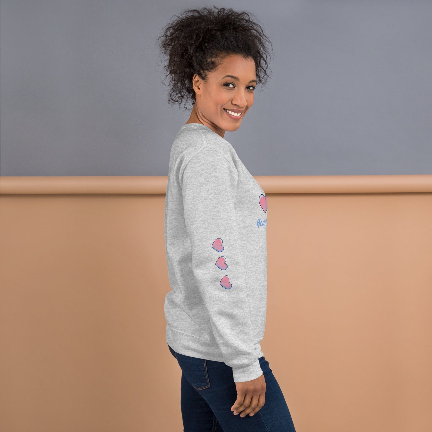 Smiling woman wearing a cozy light gray sweatshirt with three pink hearts running down the sleeve – side view showcasing playful and inspirational mom-style loungewear.