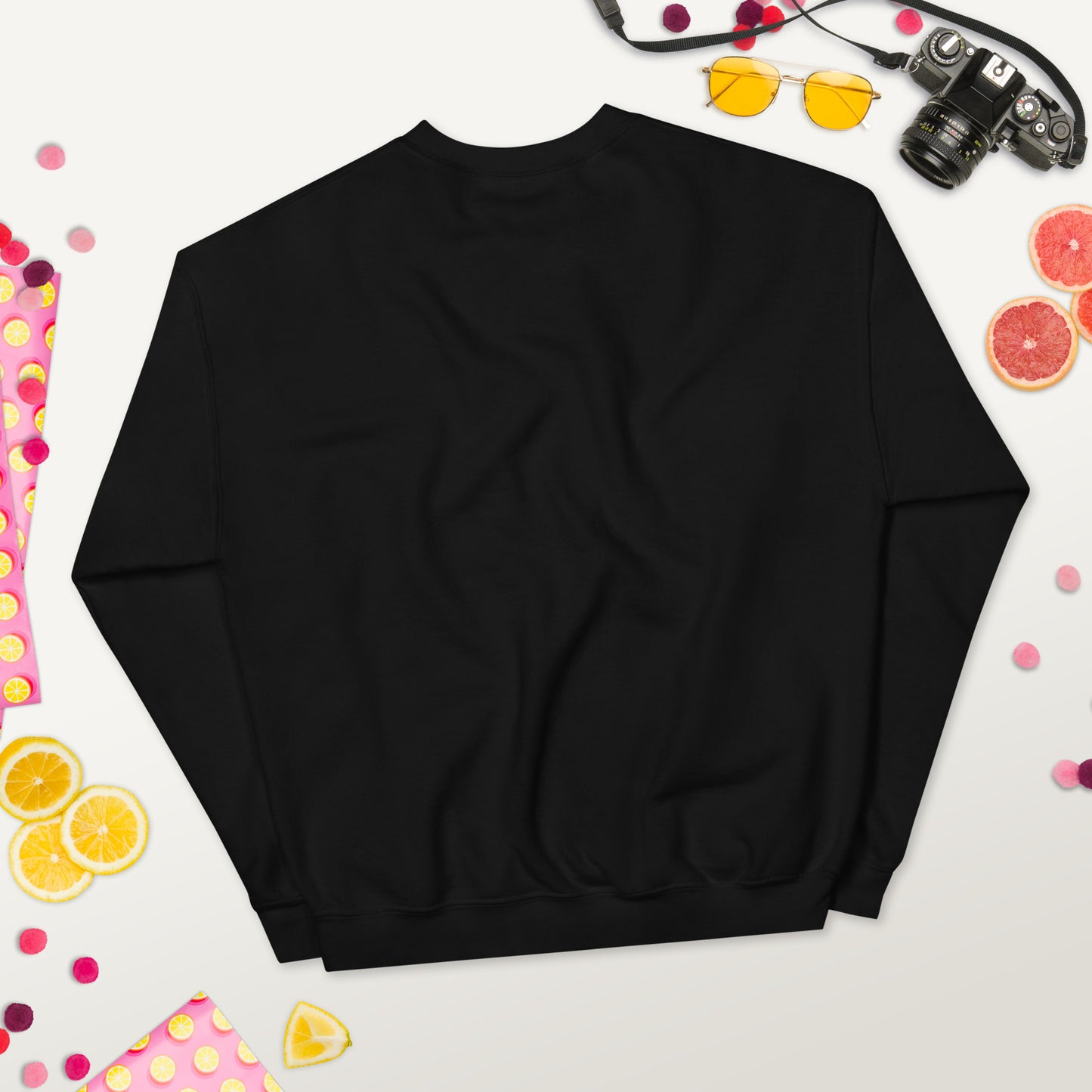 Back view of a black unisex crew neck sweatshirt laid flat on a colorful background with fruits, sunglasses, and a camera.