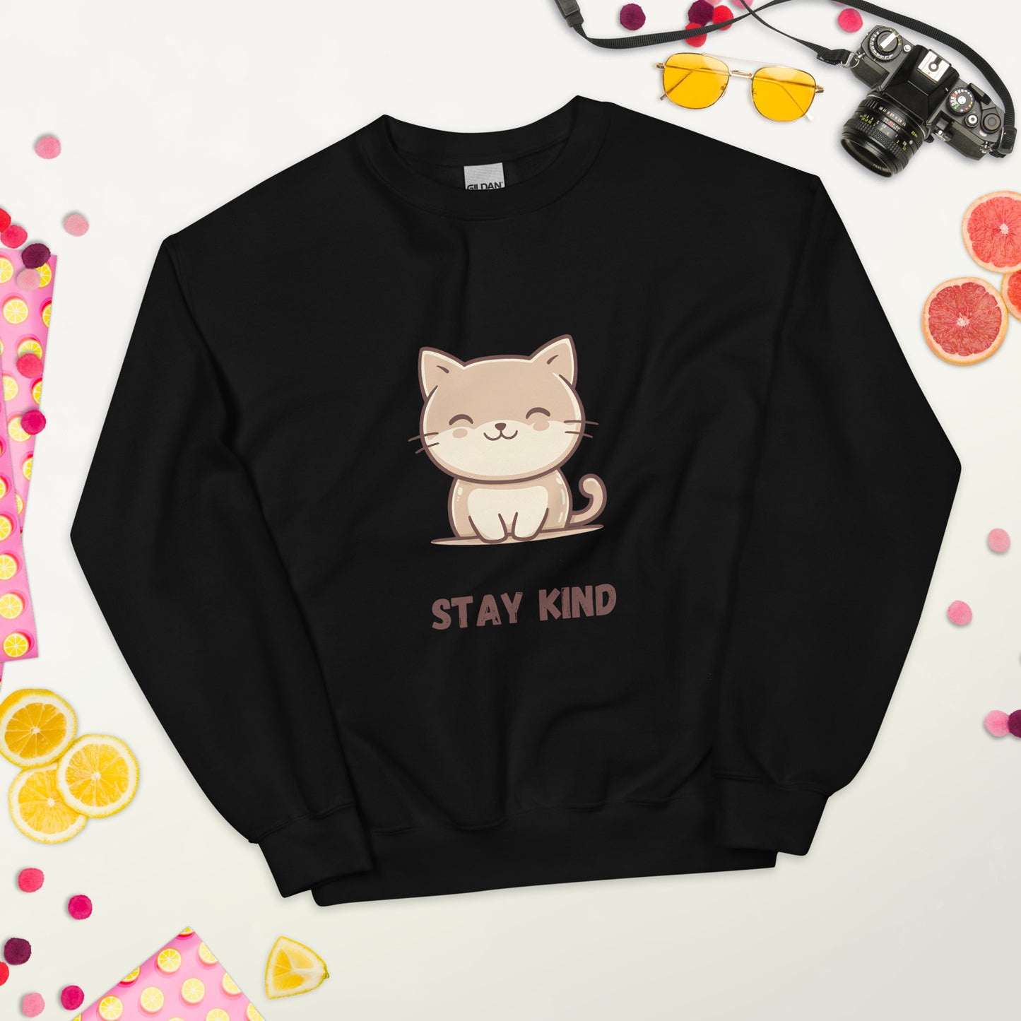 Black sweatshirt with a cute cat and the phrase “Stay Kind,” laid flat on a white surface, surrounded by colorful fruits, sunglasses, a camera, and playful decorative elements.